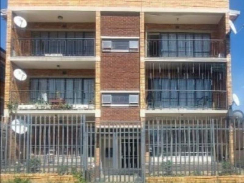 Front elevation of a multi-story brick apartment building featuring balconies, sliding glass doors, and perimeter security fencing.