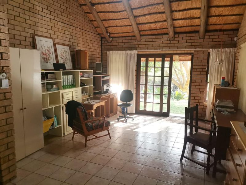 A rustic home office featuring exposed brick walls, a high thatched ceiling with timber beams, tiled floors, and direct access to the garden.