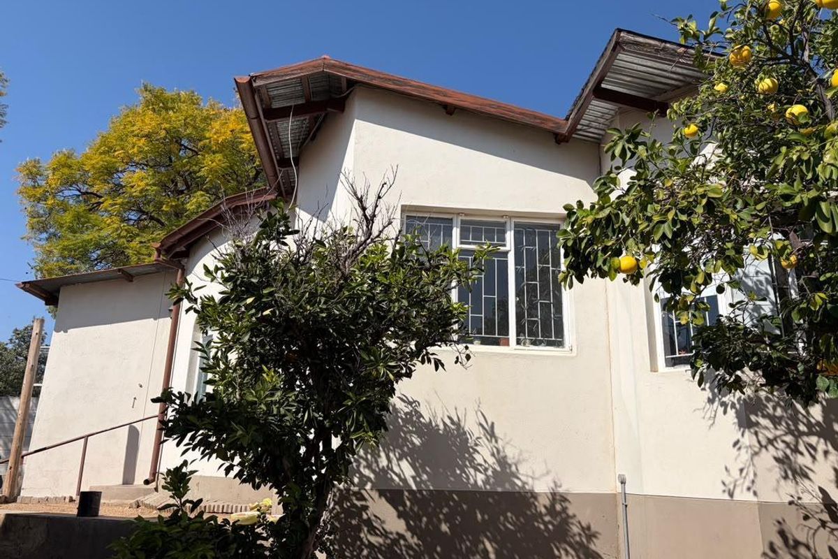 Rear exterior view of the house featuring white rendered walls, metal roofing, and security-barred windows, surrounded by mature fruit trees.