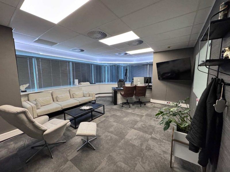 A modern, well-maintained office space featuring carpeted flooring, suspended ceilings with recessed lighting, and large windows with venetian blinds.