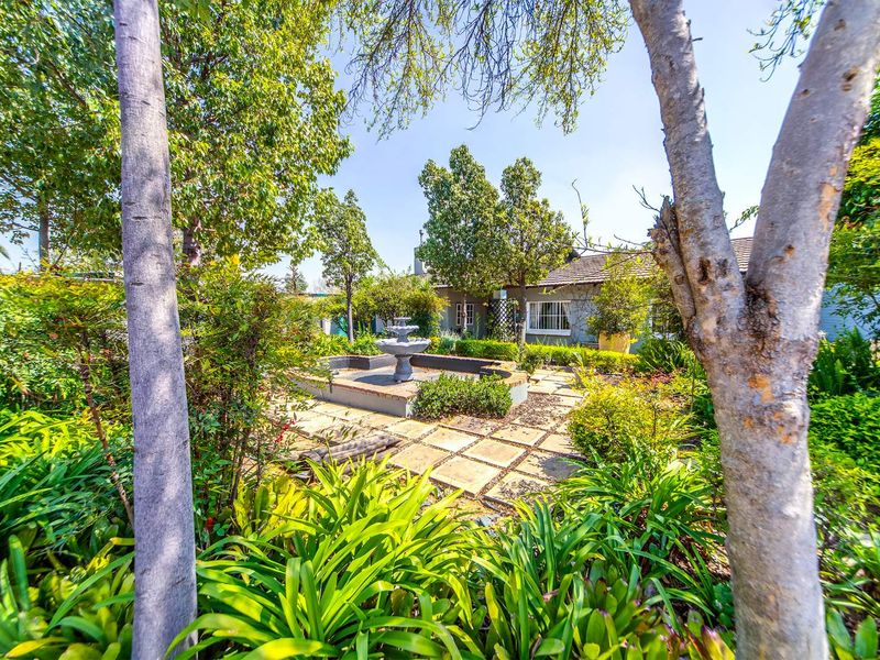A lush, well-established garden featuring a central stone fountain, paved pathways, and mature trees providing a serene outdoor setting.