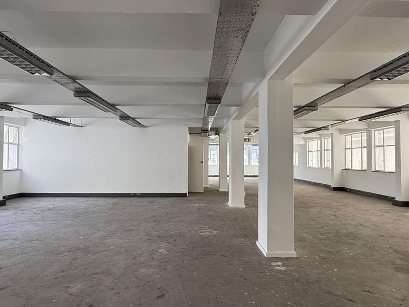 A spacious, open-plan office area featuring exposed ceiling cable trays, structural columns, and large windows providing ample natural light.