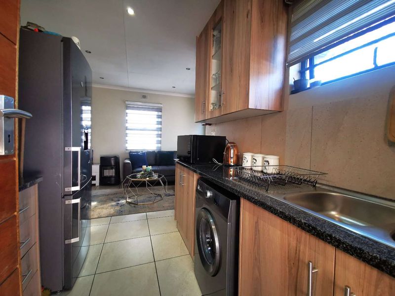 An open-plan kitchen and living area featuring modern wood-finish cabinetry, granite countertops, tiled flooring, and integrated laundry space, flowing into a comfortable lounge.