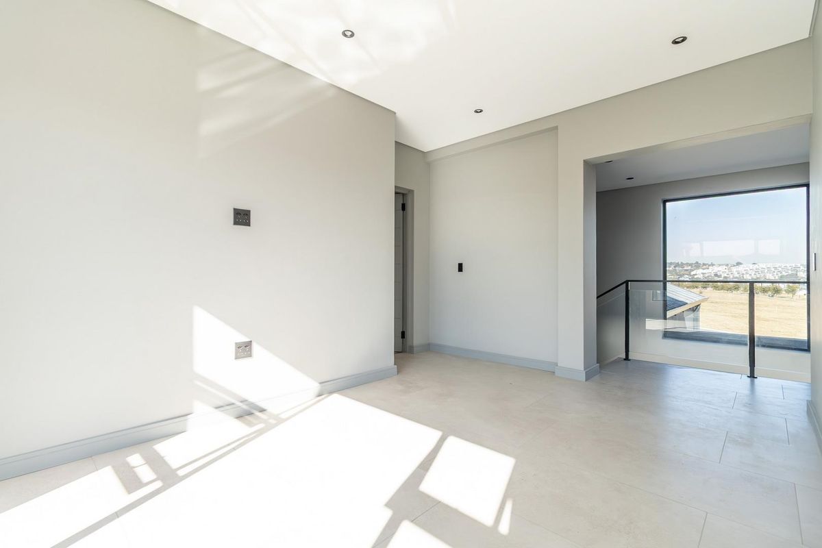 A bright, modern, and empty living space featuring neutral tiled flooring, recessed ceiling lighting, and a glass-railed staircase overlooking the surrounding landscape.