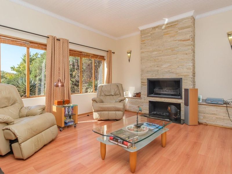A bright living room featuring a prominent stone-clad fireplace, laminate flooring, and large timber-framed windows that provide natural light and garden views.