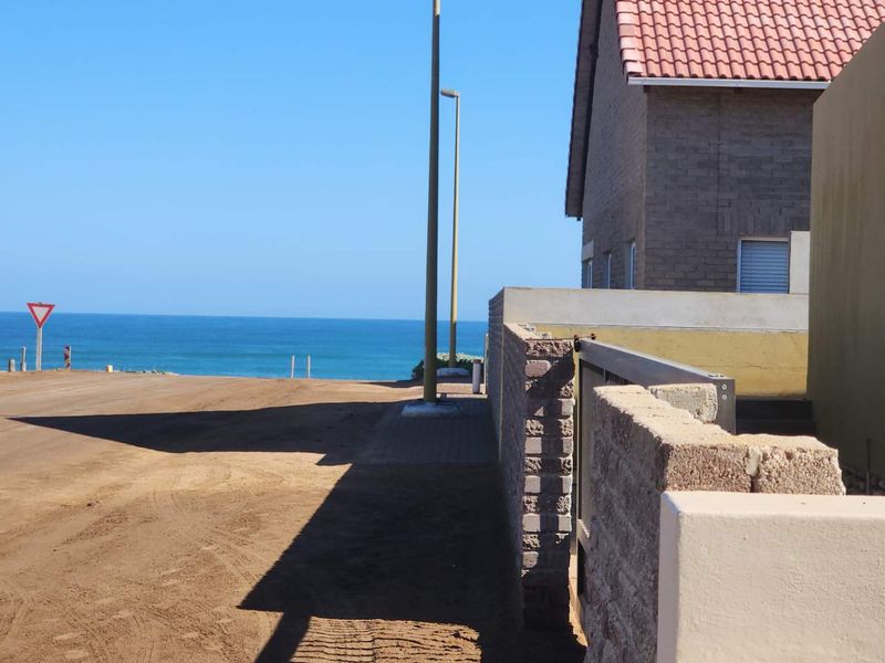 A street-level view showing the approach to the property with a brick boundary wall, tiled roof structure, and ocean views in the distance.