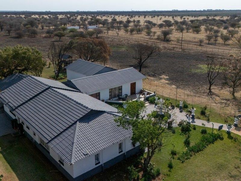 Aerial view of a farm property featuring a main dwelling with a tiled roof, paved patio area, and surrounding landscaped grounds.