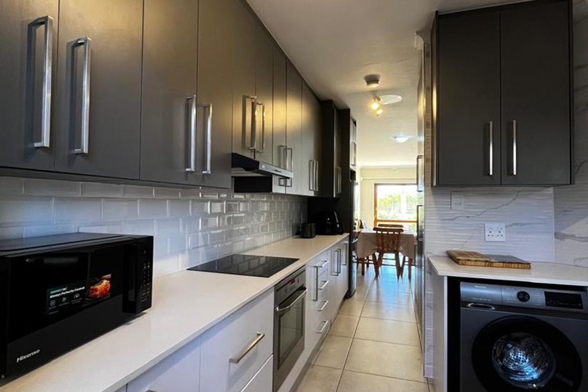 Modern galley-style kitchen featuring sleek two-tone cabinetry, white stone countertops, subway tile backsplash, integrated oven, hob, and built-in laundry appliance space.
