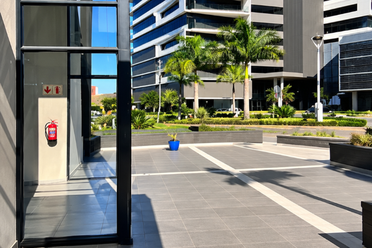 A modern paved outdoor plaza area featuring tiled flooring, glass building facade, and landscaped surroundings with palm trees in a commercial office park setting.