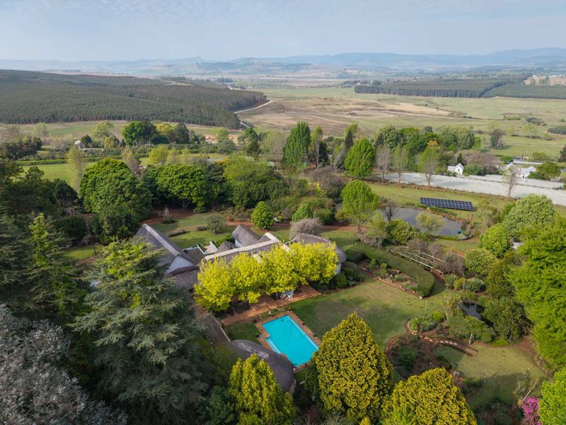 Aerial view of a farm property featuring a thatched-roof residence, swimming pool, landscaped gardens, a pond with solar panels, and expansive rural landscape views.