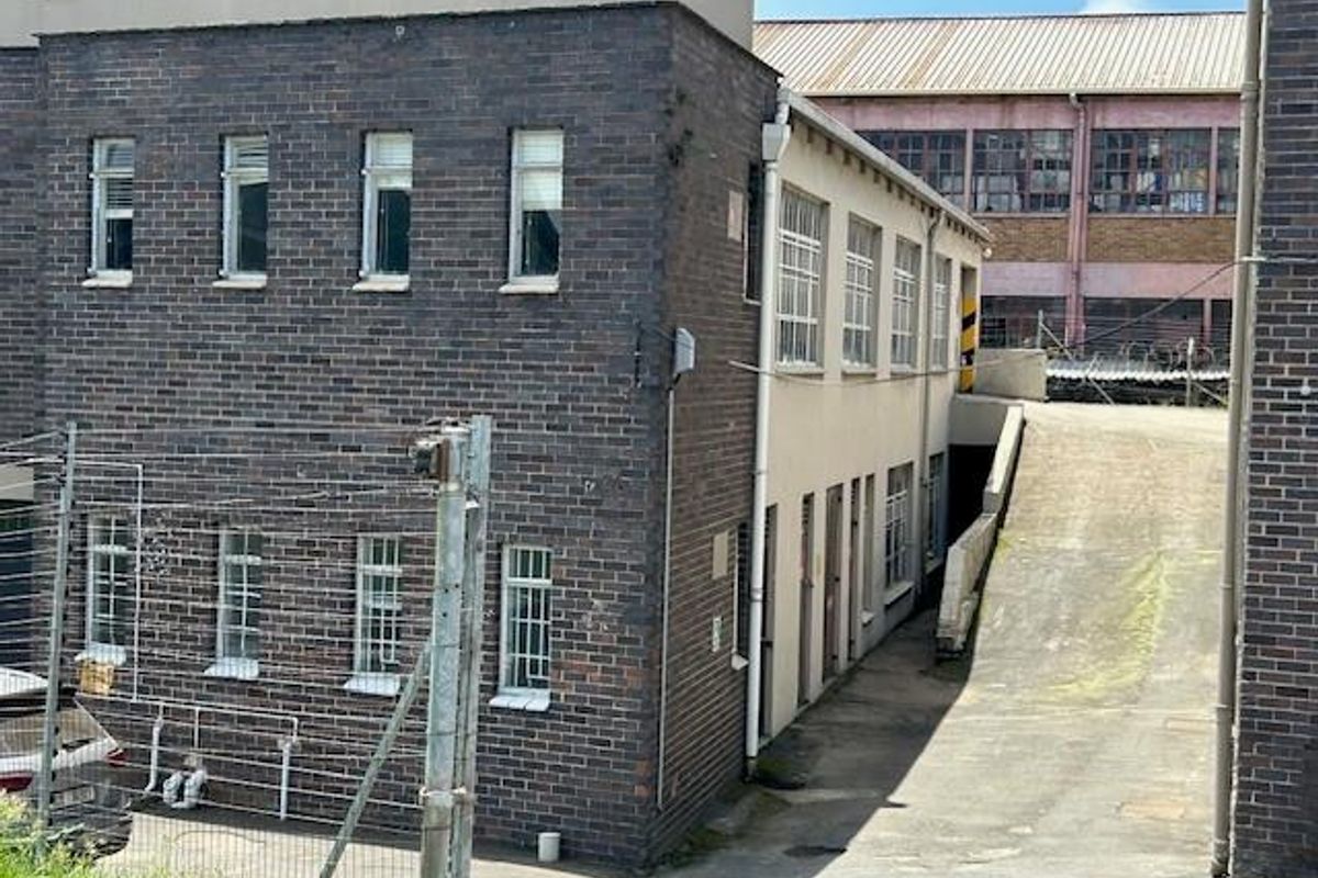 Rear exterior view of an industrial building featuring brick facade, security-barred windows, and a concrete access ramp.