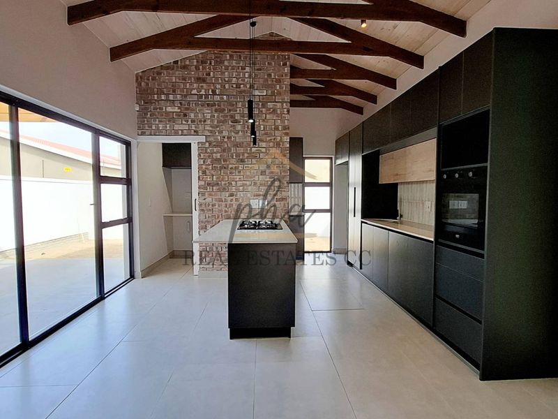Modern kitchen featuring exposed timber beams, a brick feature wall, sleek dark cabinetry, a central island with a hob, and large sliding doors.