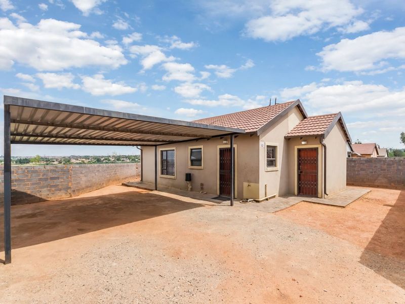 A detached single-story house featuring a large metal carport, tiled roof, and security gates, set within a fenced plot with an unpaved driveway.