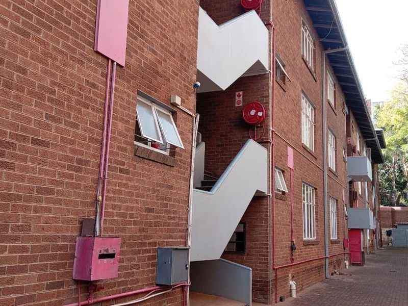 Exterior view of a multi-story brick apartment building featuring an external staircase, fire safety equipment, and a paved ground-level walkway.