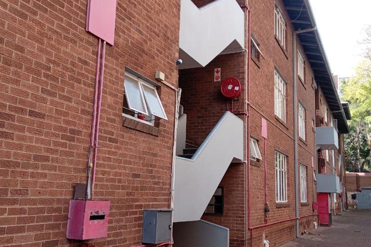 Exterior view of a multi-story brick apartment building featuring an external staircase, fire safety equipment, and a paved ground-level walkway.
