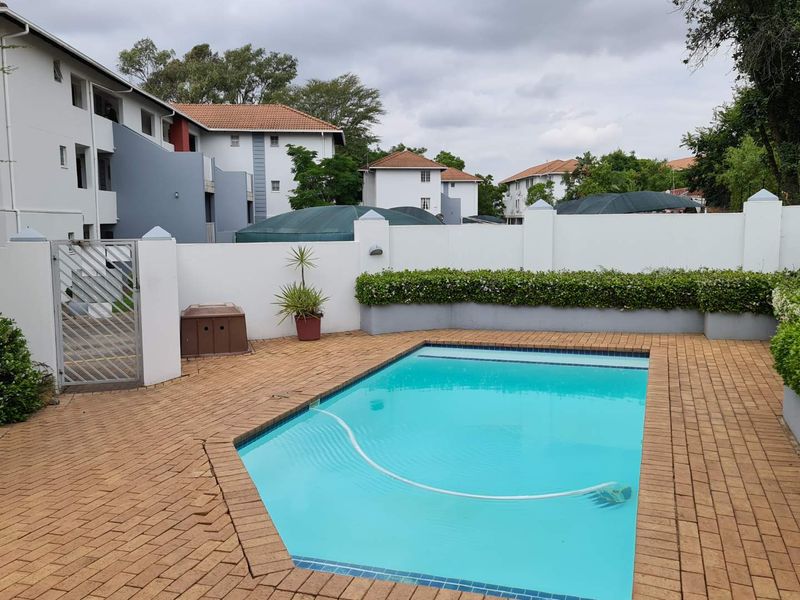A well-maintained communal swimming pool area featuring brick paving, a secure boundary wall, and established garden planter boxes.