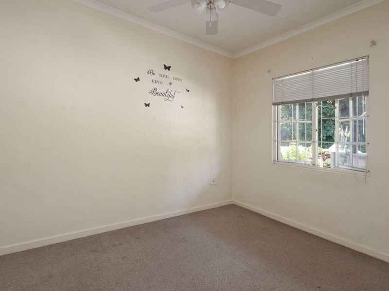A vacant bedroom featuring carpeted flooring, a ceiling fan, and a window fitted with security bars and venetian blinds.