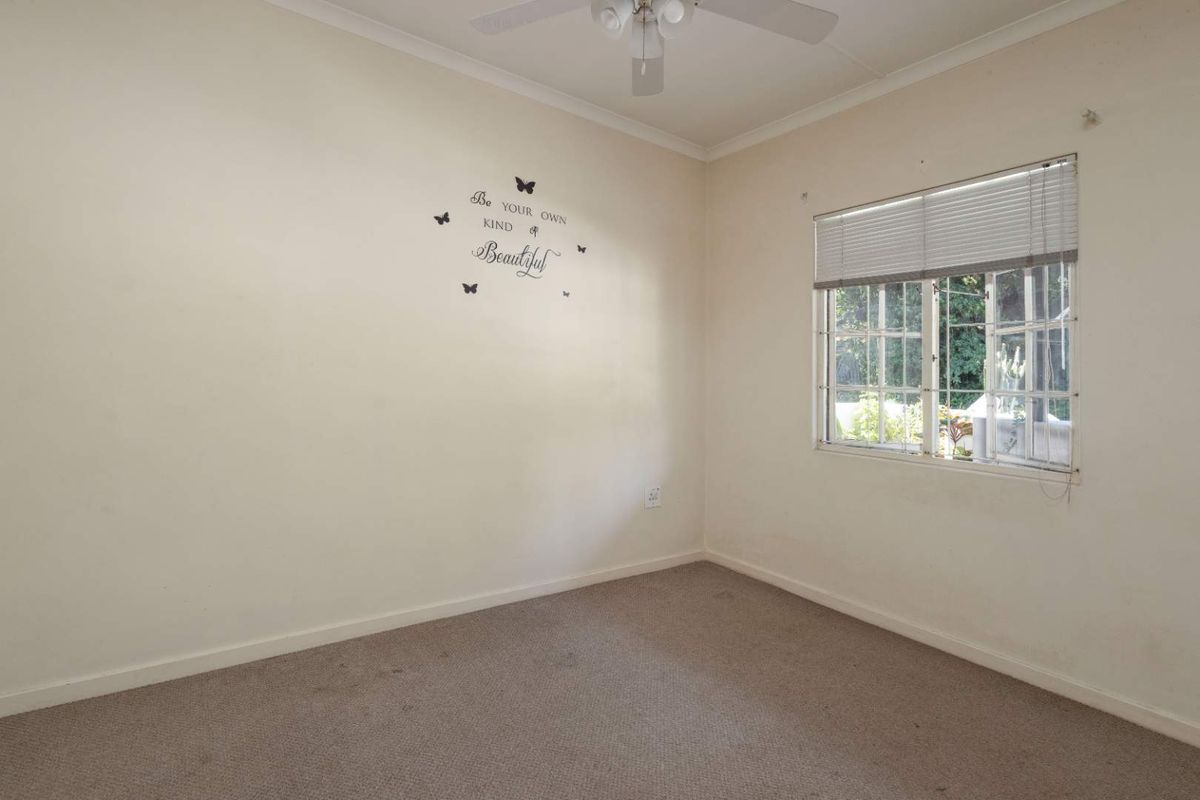 A vacant bedroom featuring carpeted flooring, a ceiling fan, and a window fitted with security bars and venetian blinds.