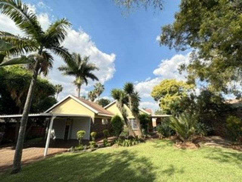 Front view of the property featuring a paved driveway, attached carport, and established garden with mature trees.
