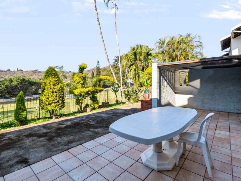 A covered patio area featuring tiled flooring, a brick support pillar, and direct access to a rear garden with scenic views.