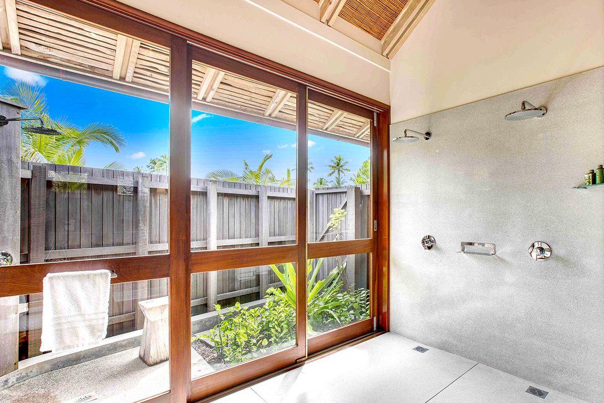 A modern, open-concept bathroom featuring dual shower heads, vaulted timber ceilings, and large glass sliding doors opening to a private outdoor garden area.