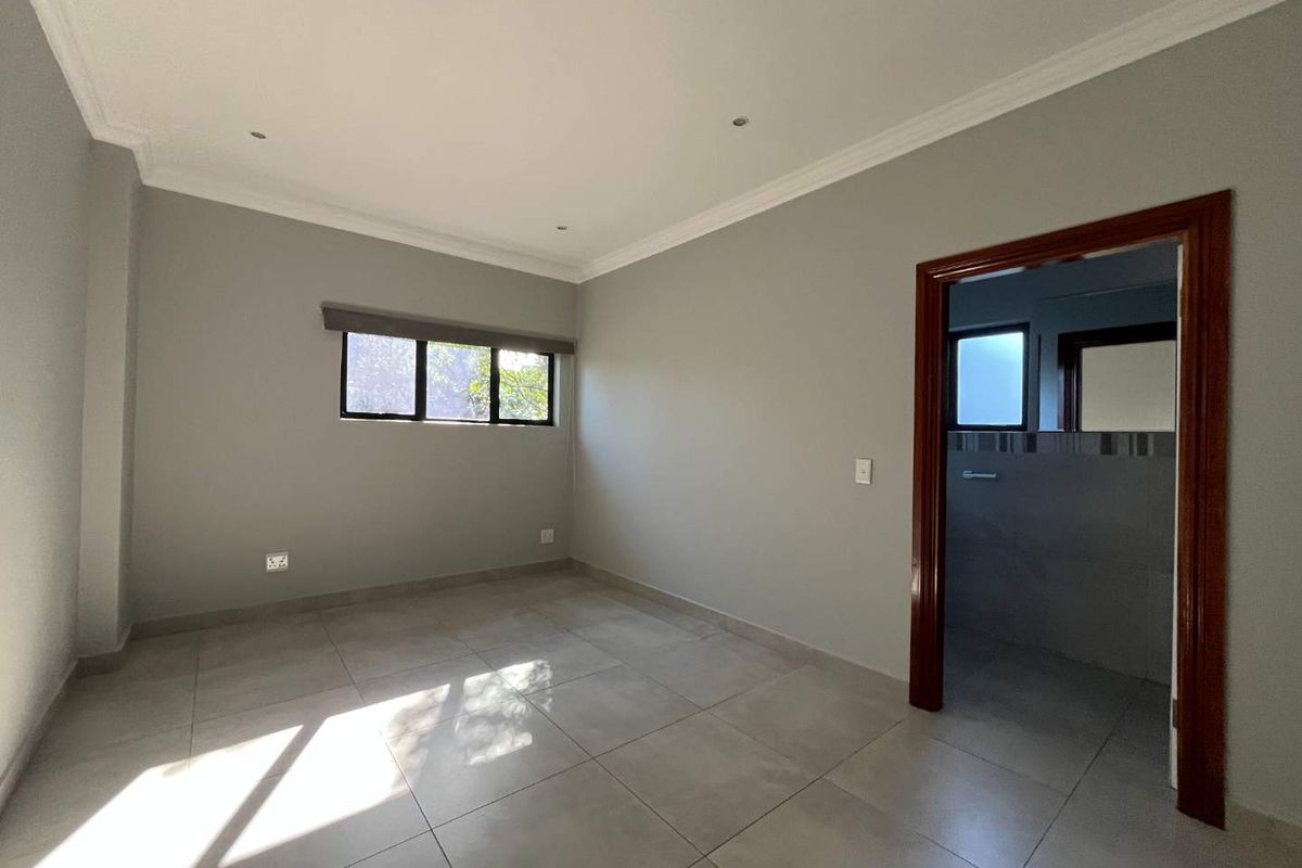 A bright, empty bedroom featuring neutral grey walls, large format floor tiles, recessed lighting, and direct access to an en-suite bathroom.
