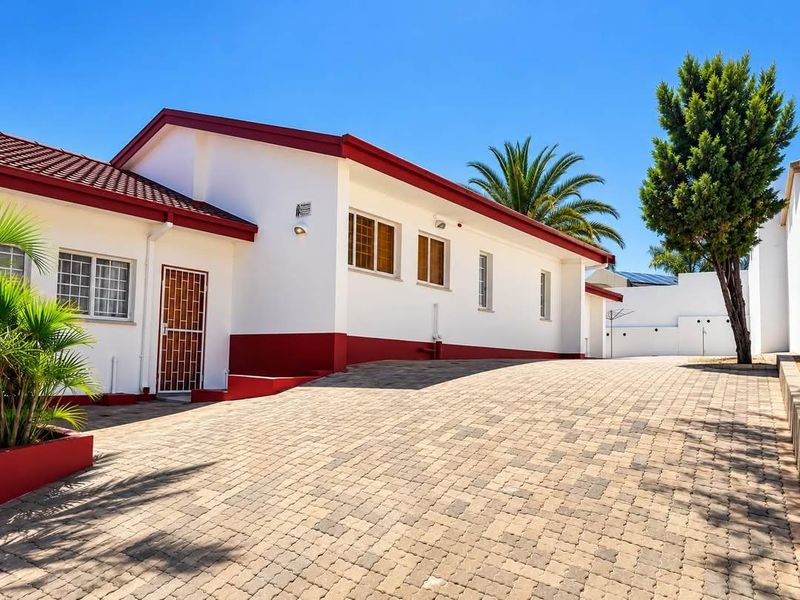 A well-maintained front exterior view of the house featuring a paved driveway, security gate, and clean white facade with red trim.