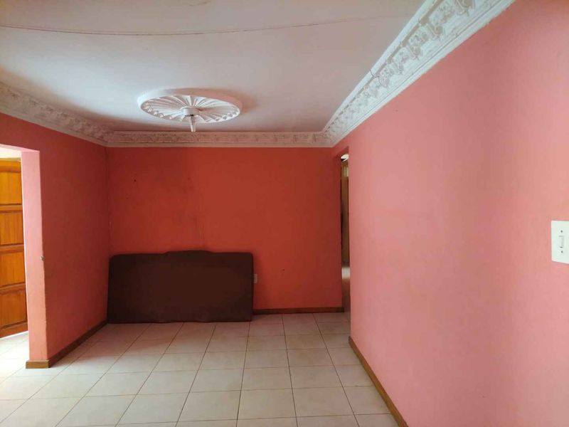 An empty living room featuring tiled flooring, decorative ceiling cornices, and a central ceiling rose, awaiting a fresh coat of paint and modern styling.