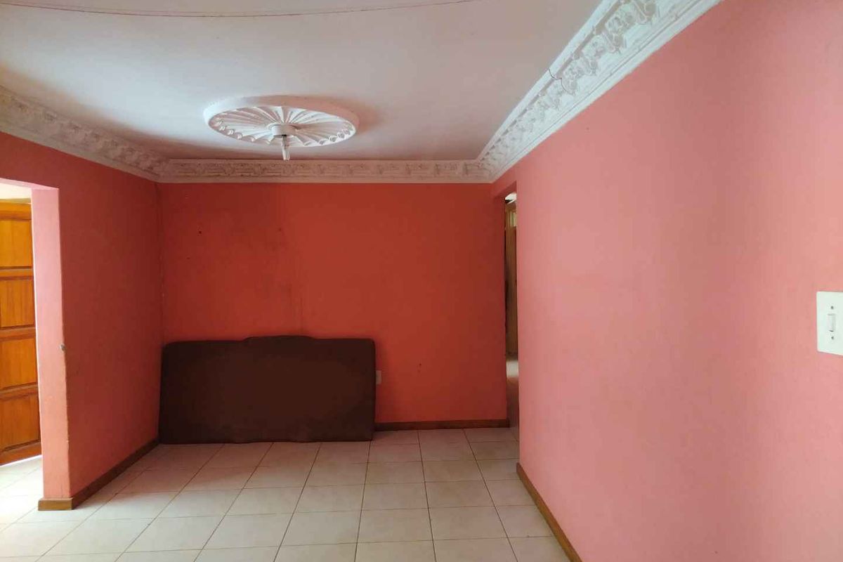 An empty living room featuring tiled flooring, decorative ceiling cornices, and a central ceiling rose, awaiting a fresh coat of paint and modern styling.
