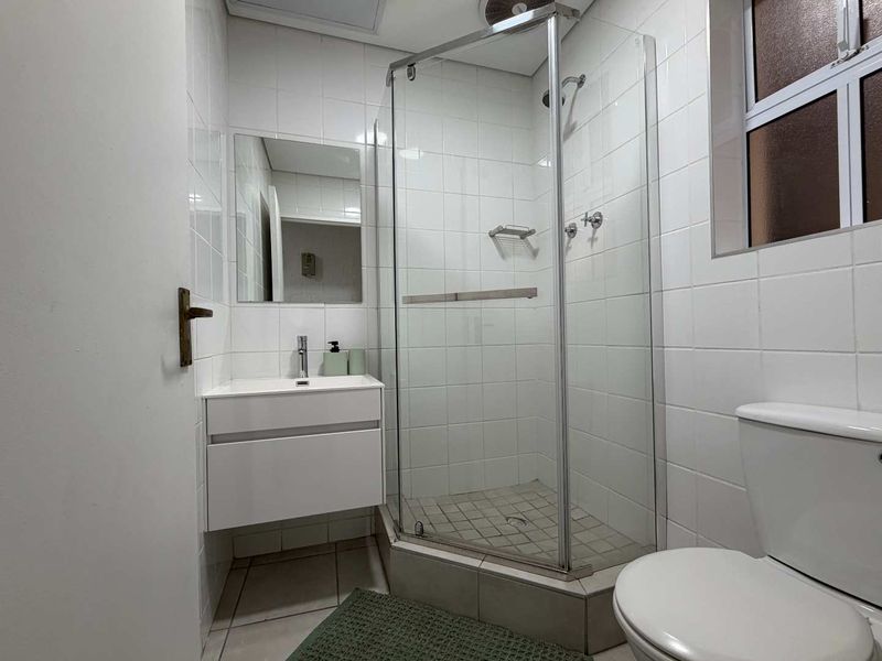 A clean, functional bathroom featuring a corner shower enclosure, wall-mounted vanity with basin, and white tiled finishes throughout.