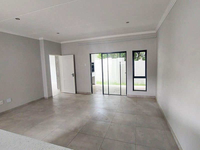 A bright, empty living area featuring neutral tiled flooring, modern aluminium-framed windows, and a sliding door providing access to the outdoor space.