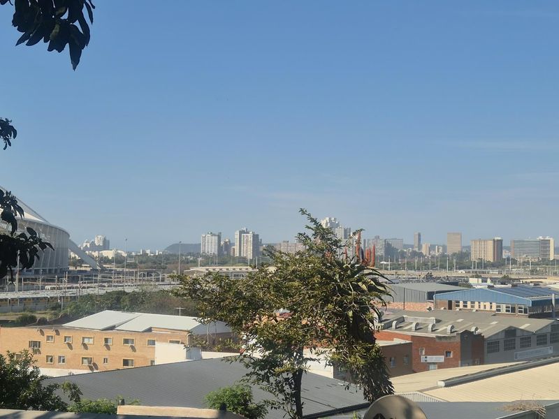 An elevated urban view overlooking the city skyline, industrial buildings, and a large stadium structure in the distance.