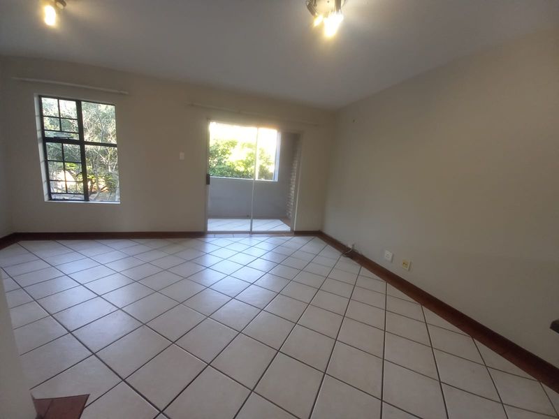 An empty, open-plan living area featuring neutral tiled flooring, natural light from a sliding glass door, and a window with security bars.