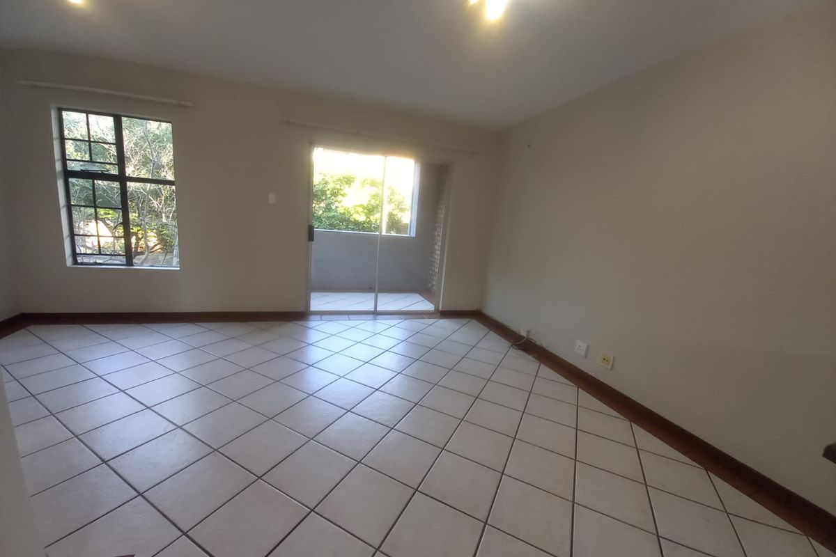An empty, open-plan living area featuring neutral tiled flooring, natural light from a sliding glass door, and a window with security bars.