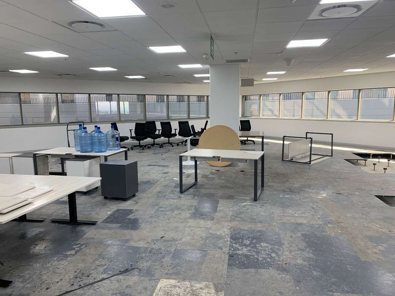 Open-plan office space featuring large windows, suspended ceilings with recessed lighting, and raised access flooring in need of repair.
