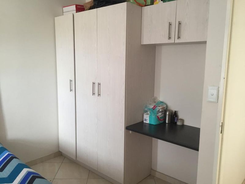 A bedroom featuring a light wood-grain built-in wardrobe with an integrated desk and overhead storage cabinets, complemented by neutral tiled flooring.