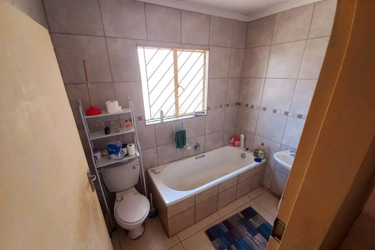 A functional bathroom featuring a built-in bathtub, pedestal sink, and toilet, finished with neutral wall and floor tiling and security bars on the window.