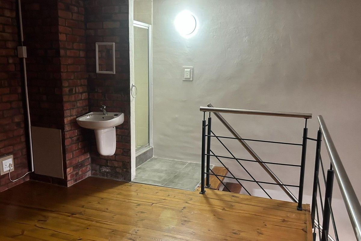 A mezzanine level bathroom area featuring exposed brickwork, a pedestal basin, and a shower enclosure, adjacent to a staircase with a modern metal balustrade.