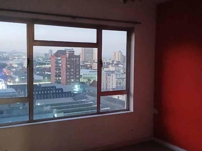 An empty bedroom featuring a large window with expansive city views and a prominent red accent wall.