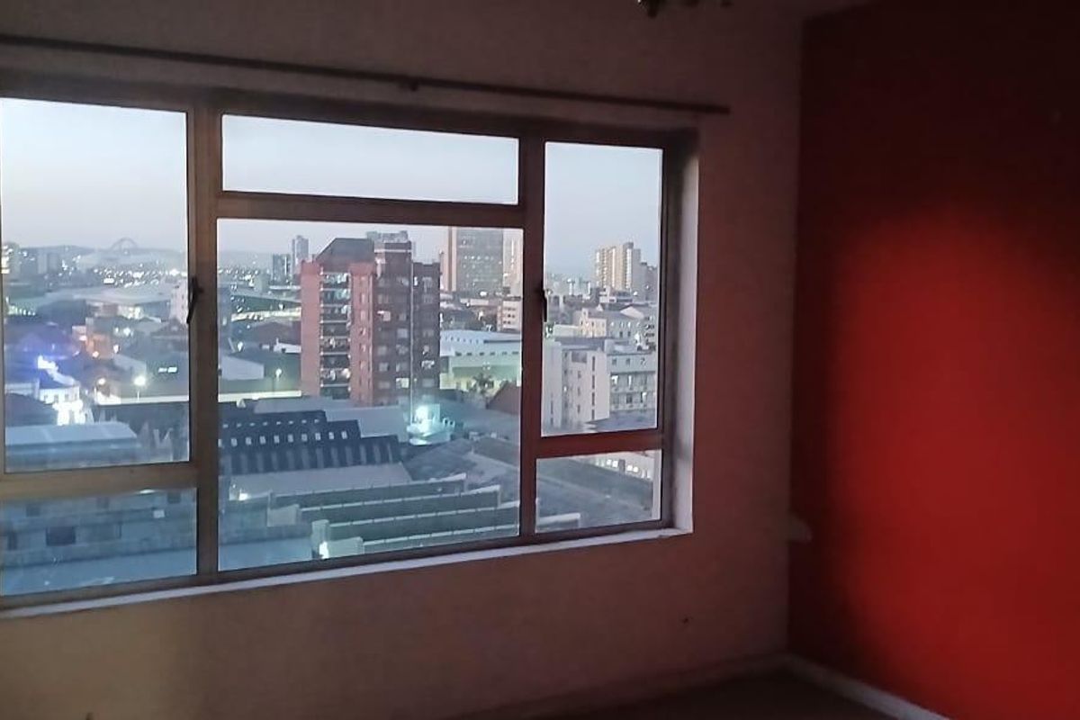 An empty bedroom featuring a large window with expansive city views and a prominent red accent wall.