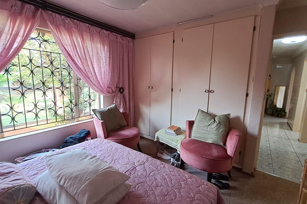 A bedroom featuring built-in wardrobes, a large window with security bars, and carpeted flooring, offering a functional space with potential for modernization.
