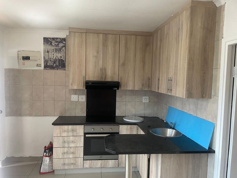 A functional kitchen featuring wood-look cabinetry, black countertops, built-in oven and hob, and a stainless steel sink with tiled splashbacks.