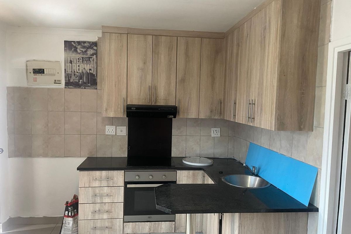 A functional kitchen featuring wood-look cabinetry, black countertops, built-in oven and hob, and a stainless steel sink with tiled splashbacks.