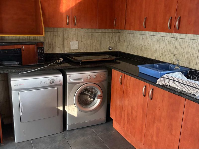 A functional laundry area featuring wood-finish cabinetry, dark granite countertops, a stainless steel sink, and tiled backsplash with integrated appliance spaces.