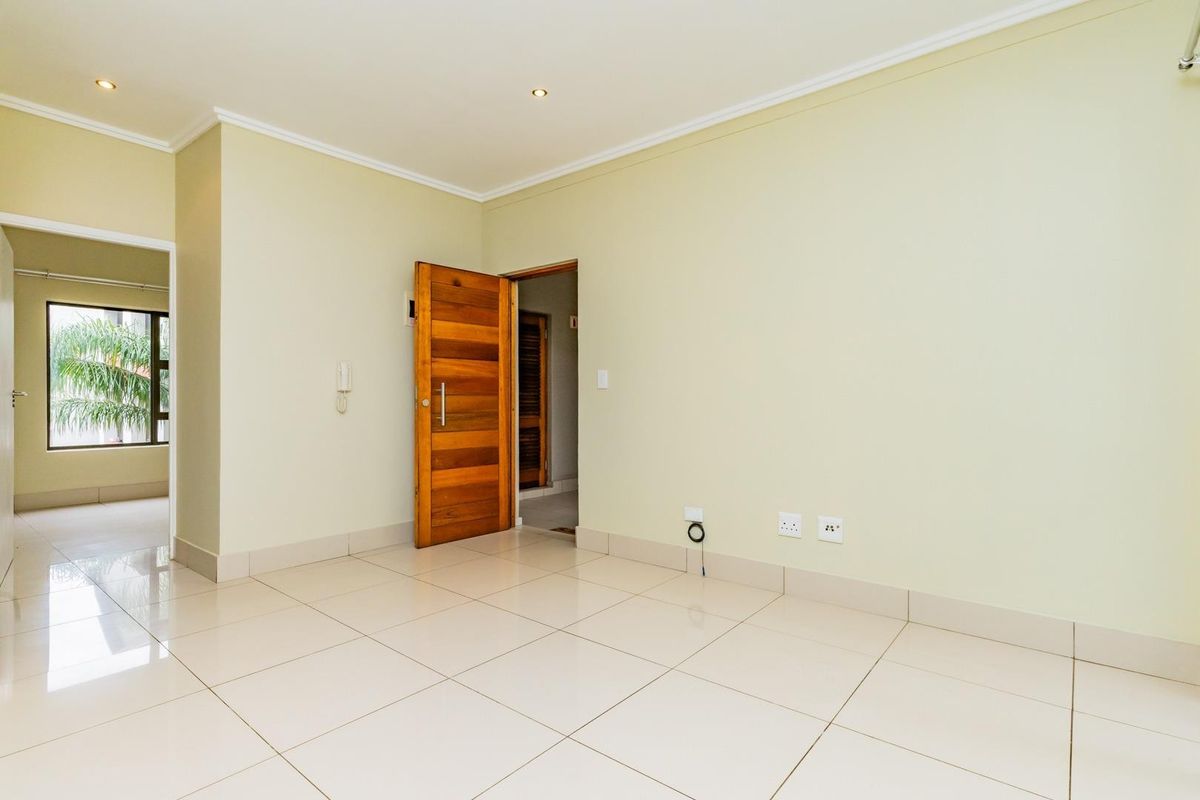 A bright, empty living area featuring neutral tiled flooring, recessed downlighting, and a solid timber door leading to an adjacent space.