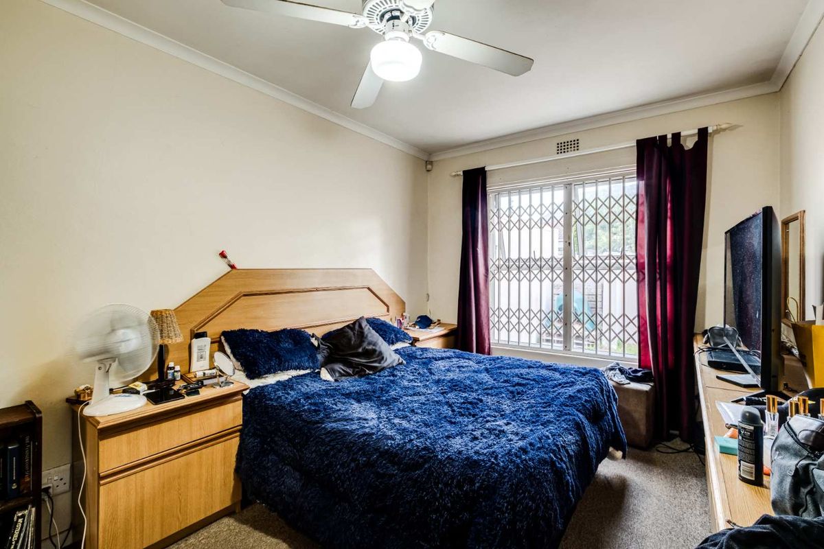 A standard bedroom featuring a ceiling fan, carpeted flooring, and a window fitted with security bars.