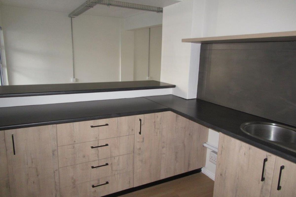 A modern, compact kitchen area featuring light wood-grain cabinetry, dark countertops, a stainless steel sink, and an industrial-style exposed cable tray.