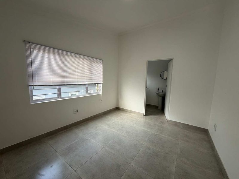 A clean, empty bedroom featuring neutral tiled flooring, a window with venetian blinds and security bars, and direct access to an en-suite bathroom.