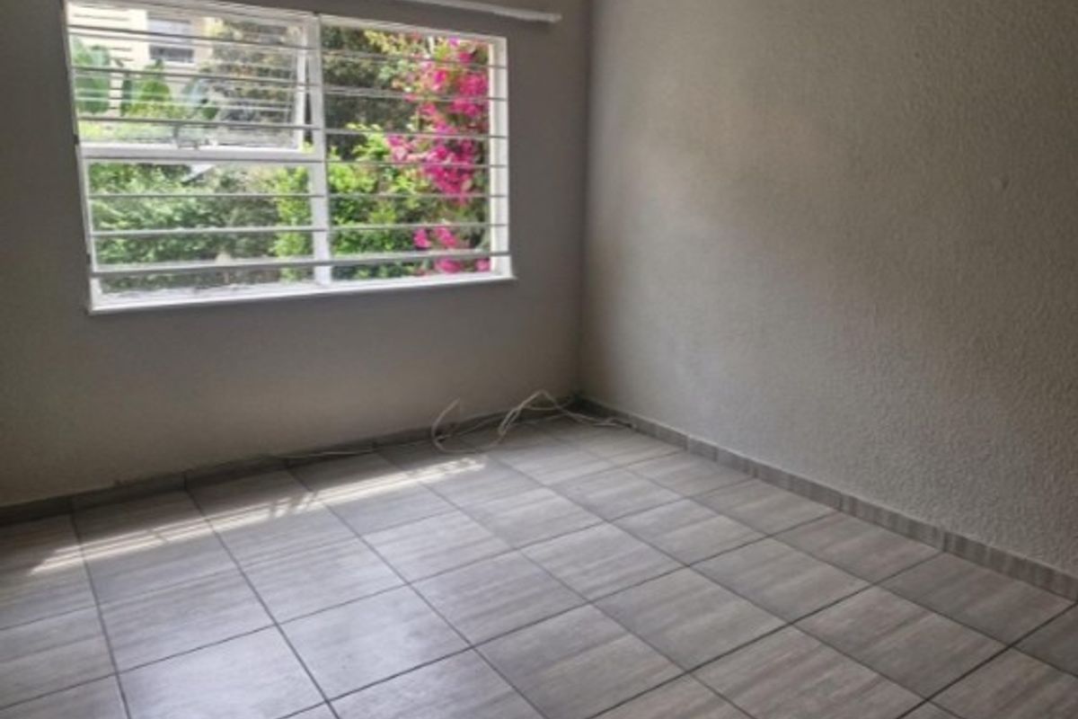 An empty bedroom featuring tiled flooring and a window fitted with security bars, offering a view of the garden.