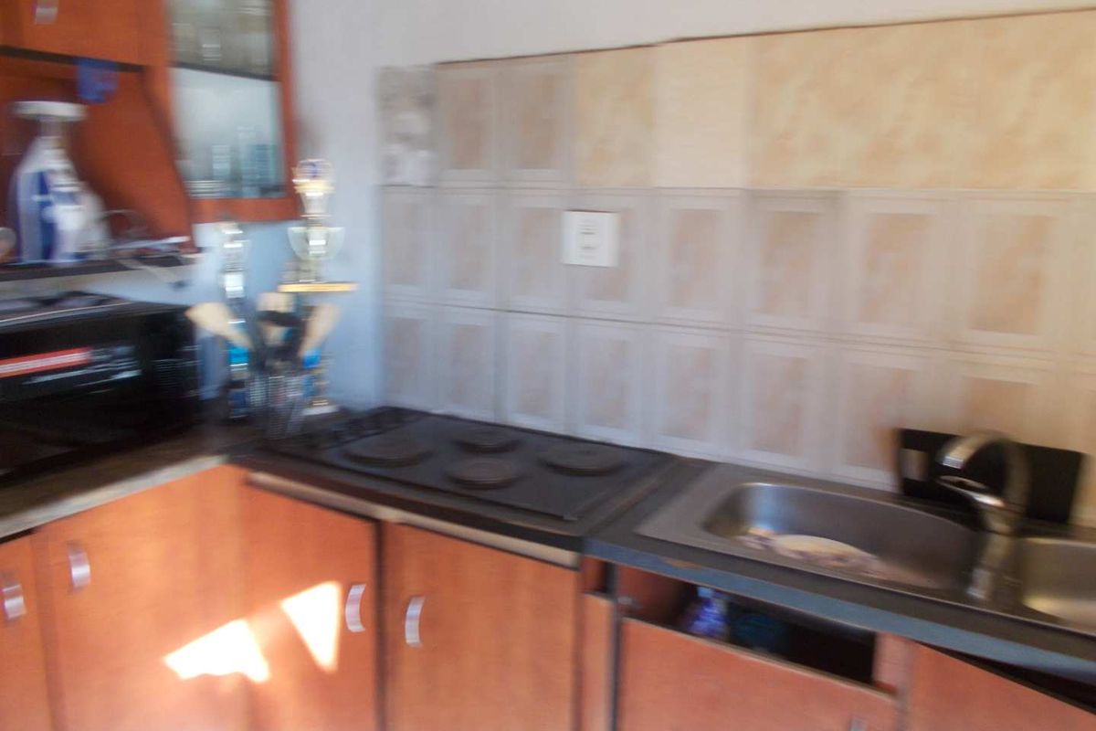 A compact kitchen featuring wooden cabinetry, a built-in electric hob, and a stainless steel sink with tiled splashbacks.